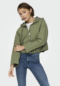 Veste cropped vert olive avec fermeture éclair et capuche, dotée de manches larges et d'un tissu léger, associée à un jean bleu foncé.
