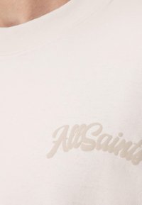 Κοντινό πλάνο ενός λευκού t-shirt με μπεζ κείμενο "AllSaints" τυπωμένο κοντά στο στήθος πάνω στο ύφασμα.