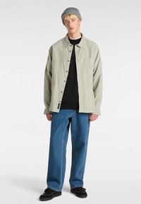 Giacca camicia in corduroy verde chiaro con maniche lunghe e bottoni, indossata sopra una camicia nera, abbinata a jeans dritti blu e scarpe nere.