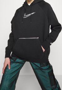 Svart huvtröja med en vit Nike-logotyp, har en framficka med dragkedja och ribbade muddar. Bärs med gröna byxor som visar en strukturerad design.