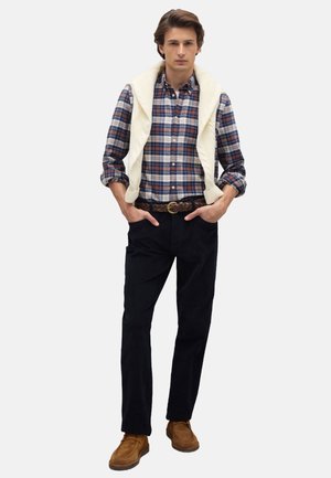 Model met een geruit overhemd in blauw, rood en wit, gecombineerd met een crèmekleurige kabeltrui, donkere broek en tankleurige veterschoenen.