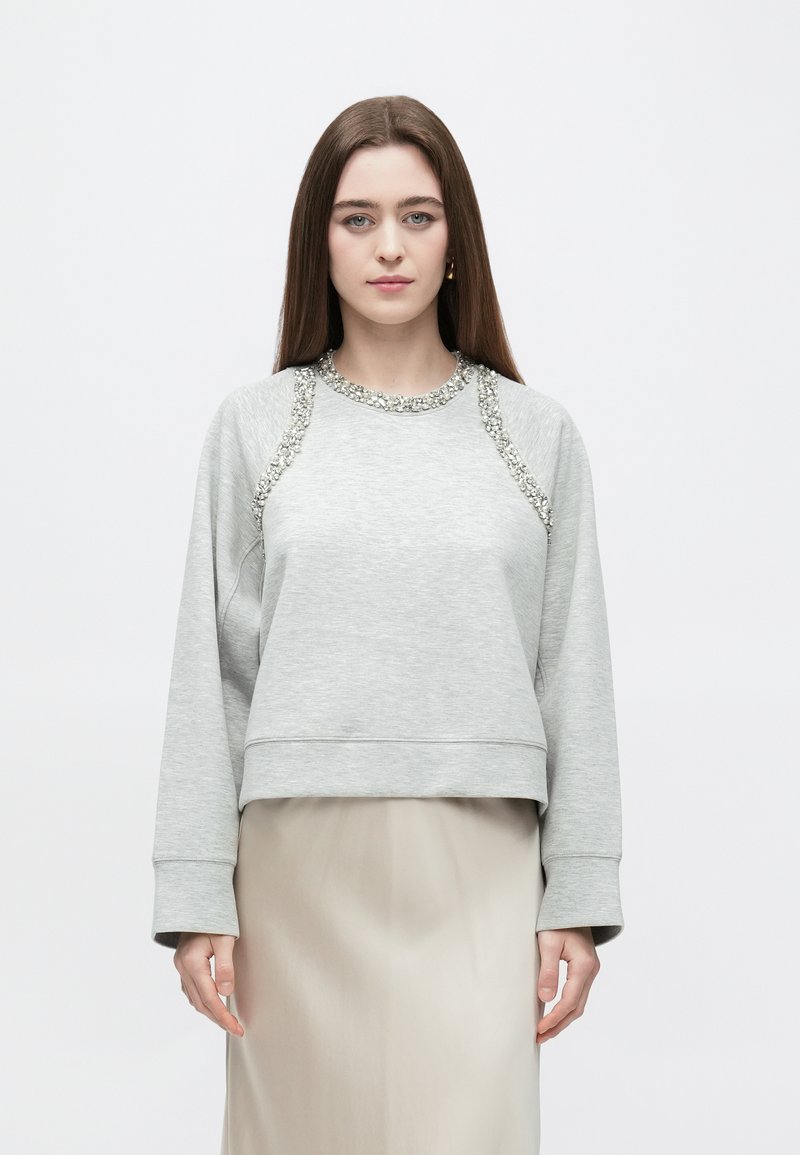 Camisola cinza clara com gola redonda, decorada com um acabamento decorativo ao longo da linha do decote e dos ombros; possui mangas compridas e um corte descontraído.