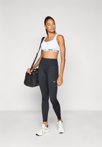 Αθλητικό σουτιέν Nike σε λευκό με λωρίδα λογότυπου, μαύρο κολάν με ψηλή μέση και λευκά αθλητικά παπούτσια· μαύρη τσάντα γυμναστηρίου με λουράκι ώμου.