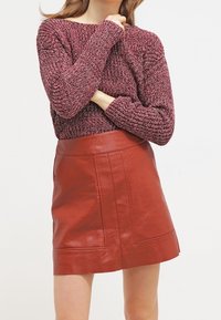 Nainen pukeutuneena viininpunaiseen neulepaitaan ja korkeavyötäröiseen ruskeaan nahkaan minihameeseen, seisomassa käsivarret ristissä yksiväristä taustaa vasten.