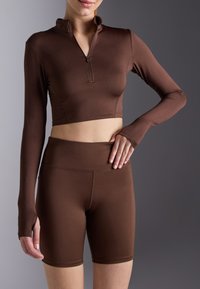 Smeđi dugi rukav uski crop top s ovratnikom na zip, uparen s odgovarajućim čistim kratkim hlačama visokog struka. Glatka, rastezljiva tkanina s prilagođenim dizajnom.