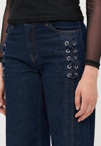 Mørke denimjeans med sidesnøring med sølvøyer og marineblå snorer, rett snitt og kontrasterende gule sømdetaljer.
