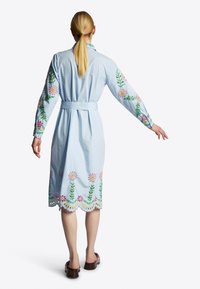 Abito camicia azzurro chiaro con ricami floreali in rosa e verde su maniche e orlo, con vita a cintura e bordi ondulati.