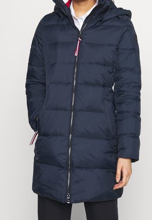 Femme portant un manteau matelassé bleu marine avec une fermeture éclair frontale, une capuche et les mains dans les poches.