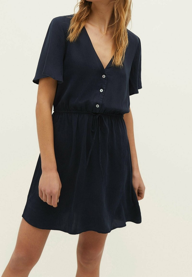 Femme portant une robe bleu marine à manches courtes avec des boutons et une taille nouée, debout devant un fond clair uni.