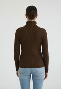 Brązowy, prążkowany sweter z golfem o dopasowanym kroju i długich rękawach, zestawiony z jasnoniebieskimi, wysokim stanem jeansami na jednolitym tle.