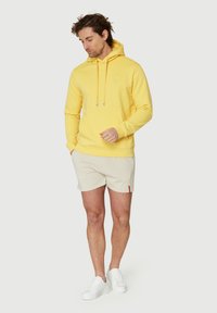 Gul hoodie med en frontlomme og snore, parret med beige shorts og hvide sneakers. Glat stof, afslappet pasform, minimal logo-detalle.