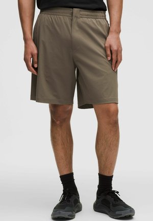 Homme portant un short à taille élastique vert olive, des chaussettes noires et des chaussures de sport noires, debout devant un fond uni.