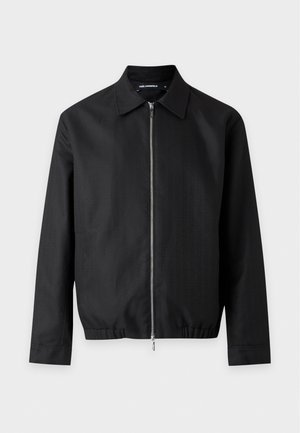 Un homme porte une veste noire zippée et un pantalon, avec une chemise blanche et des mocassins noirs et blancs, sur un fond clair et uni.