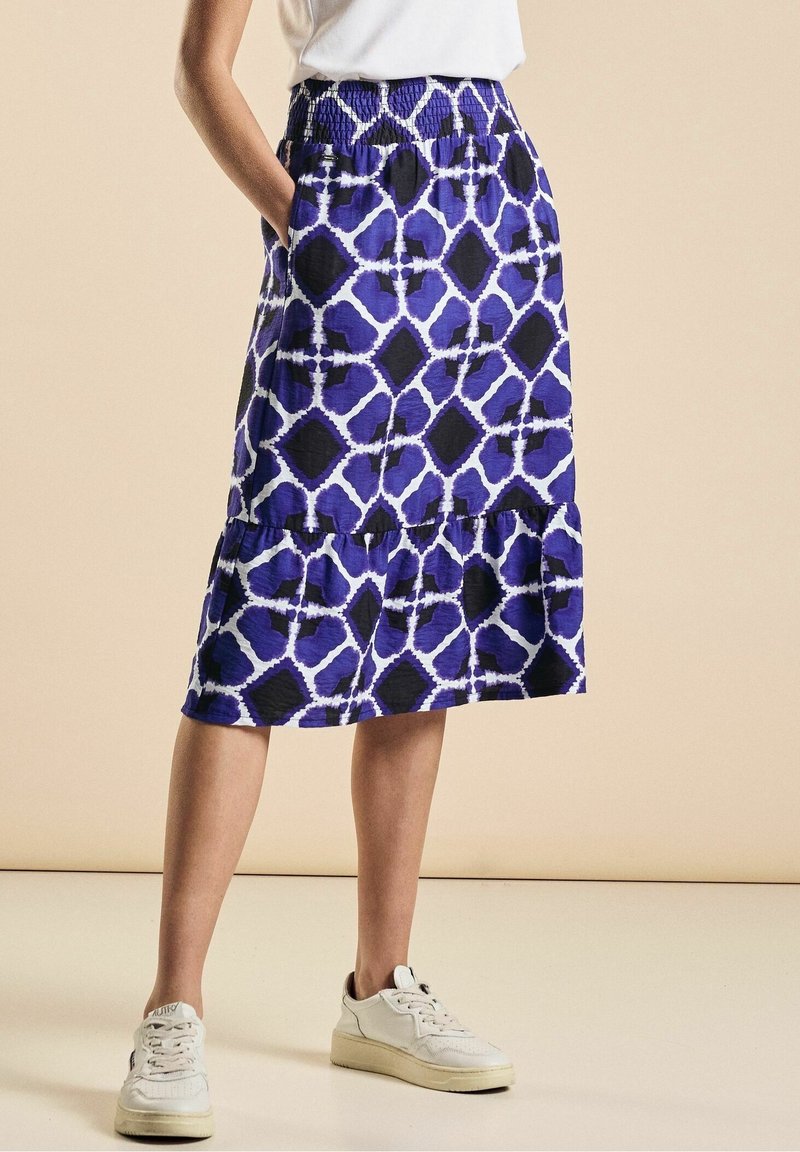 Sininen ja violetti kuvioitu hame, jossa on joustava vyötärö, kerroksellinen muotoilu ja sivutaskut, yhdistettynä valkoisiin tennareihin.