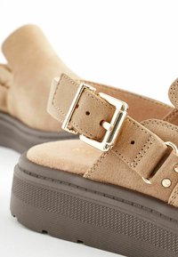 Des sandales en suède beige présentent une large sangle avec une boucle dorée, une semelle épaisse en plateforme marron et des détails de couture pour ajouter de la texture.