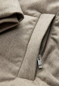 Grau-braune, weiche Stoffjacke mit einem Reißverschlussdetail. Sichtbare Nähte und Struktur; Reißverschlussanhänger mit Branding. Gefalteter Kragen nahe dem Reißverschluss.