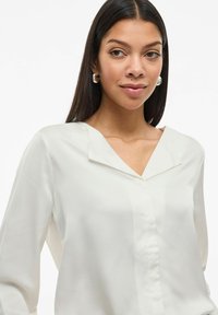 Blusa bianca con una texture morbida e liscia. Presenta un collo a V e maniche lunghe, con una leggera lucentezza e linee pulite.