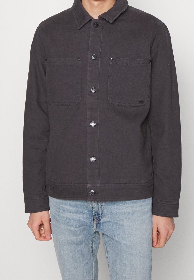 Mørkegrå jakke lavet af robust stof, med spidse kraver, lukkes foran med knapper og to brystlommer med klapper.