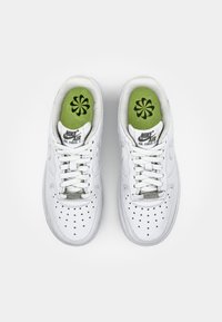 Ett par vita Nike Air Force 1-sneakers med vita snörning och gröna innersulor med ett svart blommönster, sedd uppifrån på en vit bakgrund.