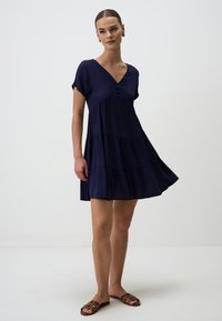 Dunkelblaues Kurzarmkleid mit V-Ausschnitt, Knopfdetails und schichtweisem Design. Der Stoff hat eine glatte Textur. Trägt dazu braune Sandalen.
