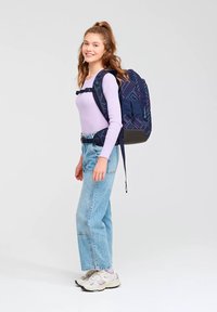 Une jeune femme souriante, en pull lavande et jeans, se tient debout avec un grand sac à dos bleu marine à motifs géométriques, ses bretelles bien attachées.