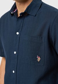 Marinblå kortärmad skjorta med knappar, tillverkad av strukturtyg, med en bröstficka på vänster sida och en färgglad broderad design.