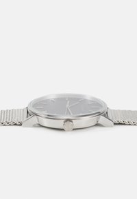 Montre en acier inoxydable avec un bracelet en maille argentée, cadran gris et marqueurs minimalistes. Présente une couronne saillante avec le logo "CK" sur le côté.