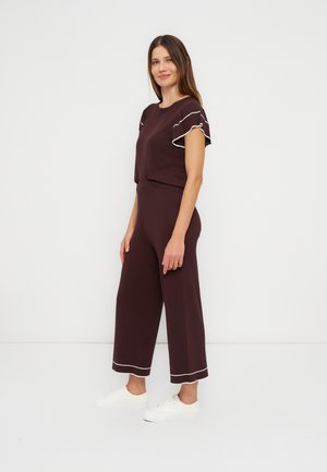 Femme debout portant une combinaison pantalon violet foncé avec des bordures blanches aux manches et aux ourlets du pantalon, associée à des baskets blanches, sur un fond uni.