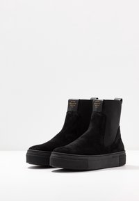 Botines de ante negros con paneles laterales elásticos, suela gruesa de plataforma y logo grabado en el tacón. Textura suave y diseño elegante.