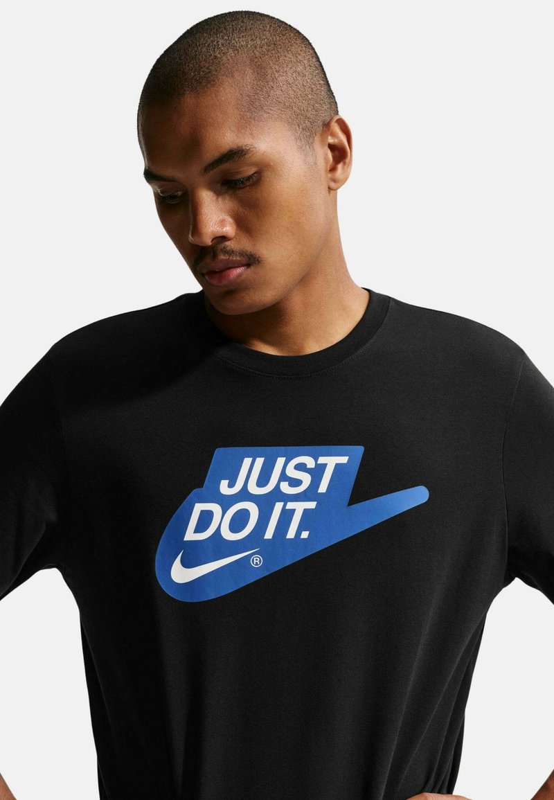 Joven con el cabello corto que lleva una camiseta negra con el logo azul de Nike y el texto "JUST DO IT." mirando hacia abajo.