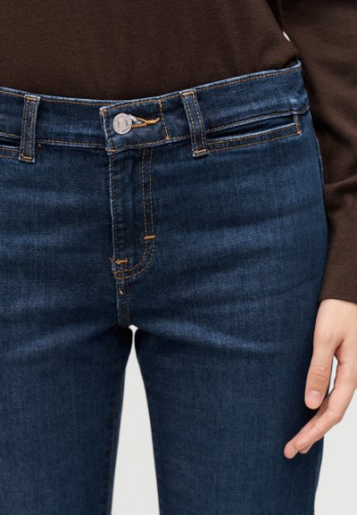 Άτομο που φορά σκούρο μπλε denim τζιν με πορτοκαλί ραφές και καφέ μακρυμάνικη μπλούζα, το χέρι ξεκουράζεται στο πλάι.