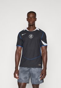 Maglietta sportiva nera con accenti blu e bianchi, caratterizzata da un collo rotondo, maniche corte e texture. Indossata con pantaloni corti grigi a pois.