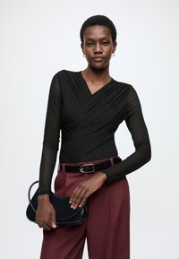 Μαύρο κορμάκι τύπου wrap με μακριά διάφανα μανίκια, συνδυασμένο με ψηλόμεσα μπορντό παντελόνια και μικρή μαύρη τσάντα χειρός. Λείο ύφασμα και εφαρμοστή γραμμή.