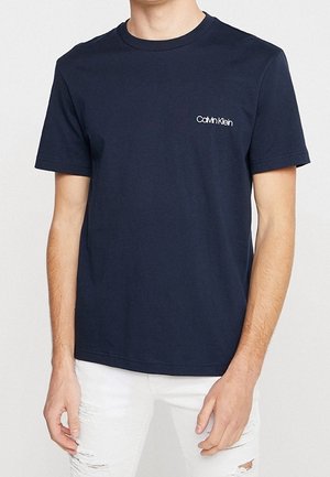Homme portant un t-shirt à manches courtes bleu marine uni avec le logo "Calvin Klein" sur la poitrine gauche et un jean blanc déchiré.