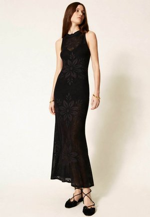 Donna che indossa un abito lungo nero lavorato all'uncinetto senza maniche con motivi floreali e ballerine nere con lacci, in piedi davanti a uno sfondo chiaro uniforme.