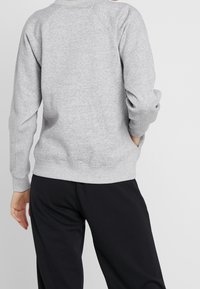 Persona vestida con un sudadera de manga larga gris claro y pantalones negros, mostrada de espaldas con una mano en el bolsillo.