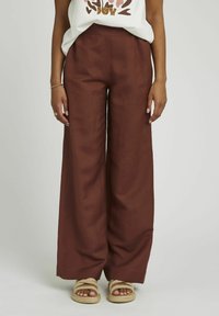 Pantalons large en lin de couleur rouille profond, avec une taille haute. Associés à des sandales tressées beiges.