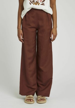 Pantalons large en lin de couleur rouille profond, avec une taille haute. Associés à des sandales tressées beiges.