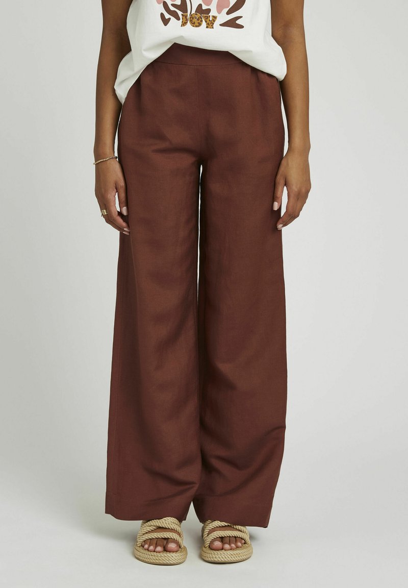 Pantalons large en lin de couleur rouille profond, avec une taille haute. Associés à des sandales tressées beiges.