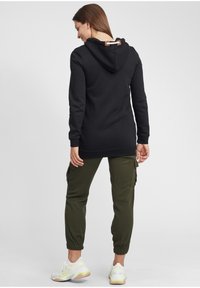 Felpa nera con cappuccio e un loop decorativo sul retro, abbinata a pantaloni cargo verde oliva e sneakers bianche con dettagli gialli.