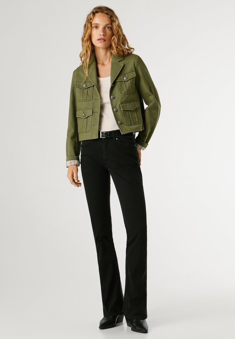 Kobieta ubrana w oliwkowo-zieloną zapinaną na guziki kurtkę, biały top, czarne spodnie z paskiem i czarne buty na obcasie, stojąca na tle jasnego, jednolitego tła.