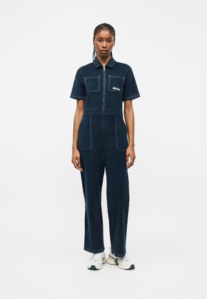 Žena stojaca, oblečená v námornícky modrom overal s krátkym rukávom na zips, s prednými vreckami a bielymi teniskami, na bielom pozadí.