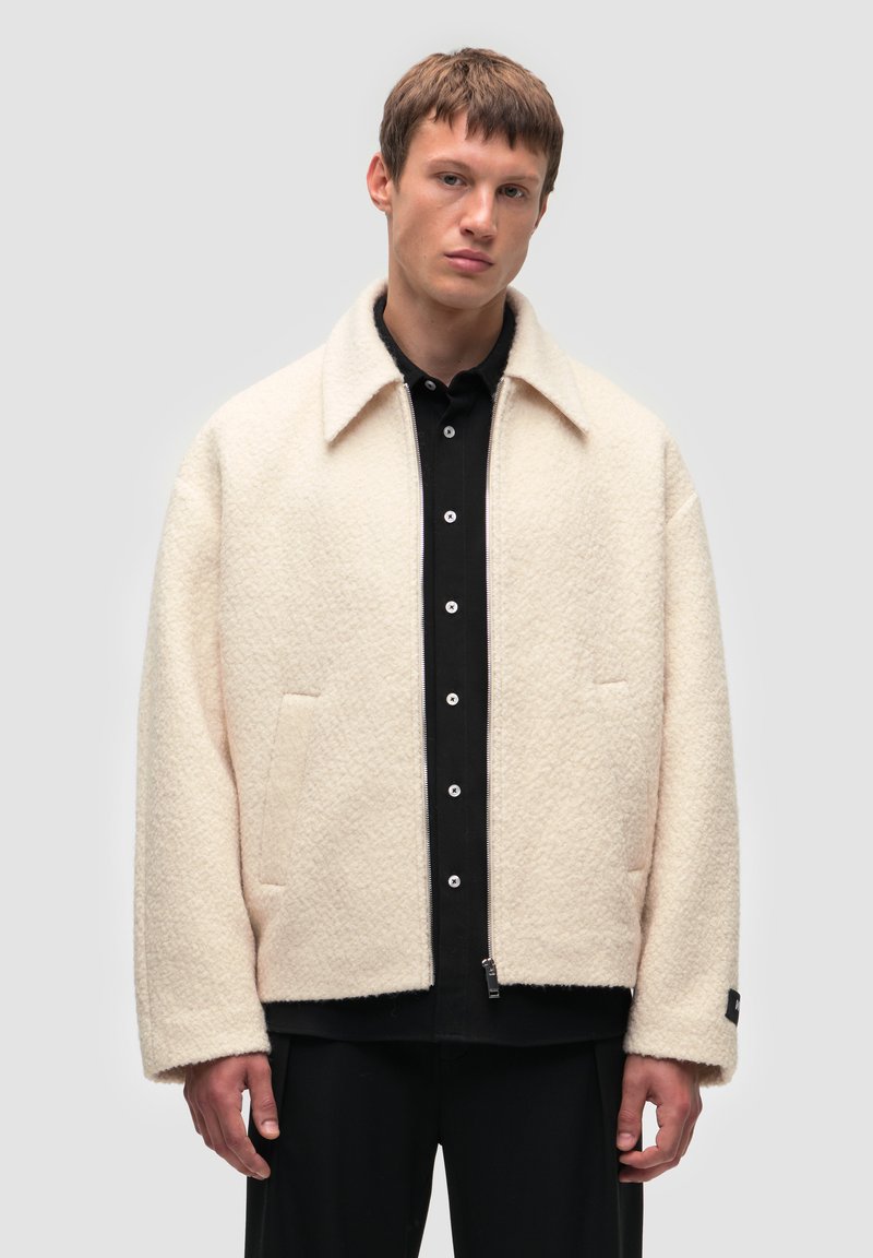 Biała, fakturowana wełniana kurtka z kołnierzykiem, dwoma przednimi kieszeniami i zapięciem na zamek, noszona na czarnej koszuli zapinanej na guziki.