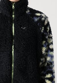Chaqueta de forro polar con cuello alto, cuerpo negro y mangas con un patrón en azul, verde y crema. Presenta una cremallera verde clara y un detalle del logo.