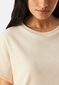 Frau mit langen dunklen Haaren, die ein strukturiertes, cremeweißes Kurzarmoberteil mit rundem Ausschnitt trägt, wobei ihr unteres Gesicht und ihr Hals sichtbar sind.