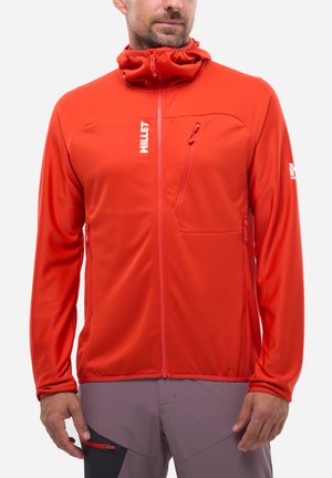 Mann trägt eine leuchtend rote Outdoor-Jacke mit Reißverschluss und Kapuze, Brust- und Seitentaschen mit Reißverschluss, sowie graue Hose mit kleiner roter Reißverschlusstasche.