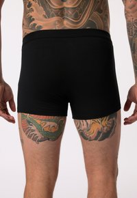 Shorts en coton noir avec une coupe ajustée et une taille élastique, ornés de tatouages détaillés sur les cuisses avec des couleurs vives et des motifs complexes.