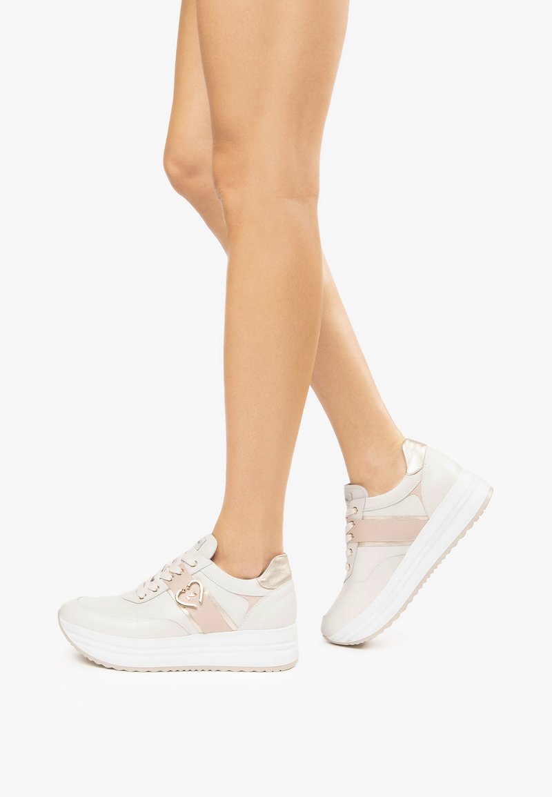 Baskets blanches et beiges avec un accent métallique, dotées d'une semelle surélevée, d'un design à lacets et d'un détail en forme de cœur sur le côté.
