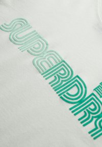 Biała bawełniana koszulka z multikolorowym, welurowym napisem "SUPERDRY" w zielonym kolorze, z nałożonym retro krojem czcionki.