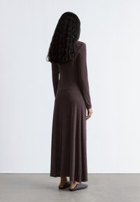 Langes braunes Kleid mit langen Ärmeln und einem fließenden Rock, das ein geprägtes Muster aufweist. Getragen mit dunklen Slippern.